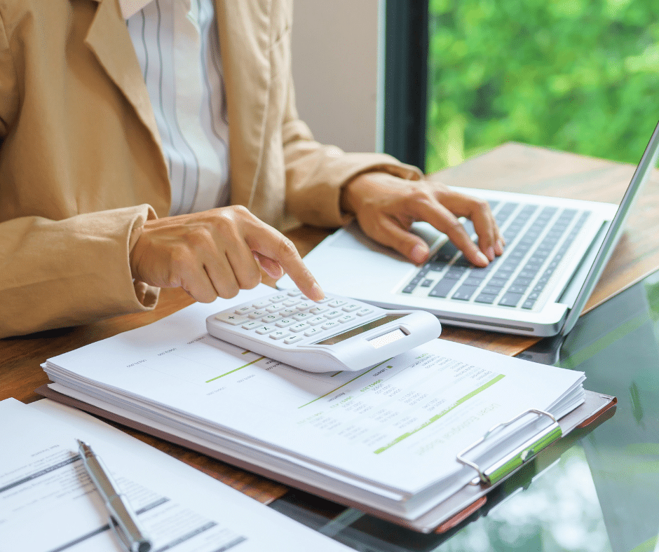 Person balances a budget using a calculator and laptop over printed financial documents on a wooden desk.