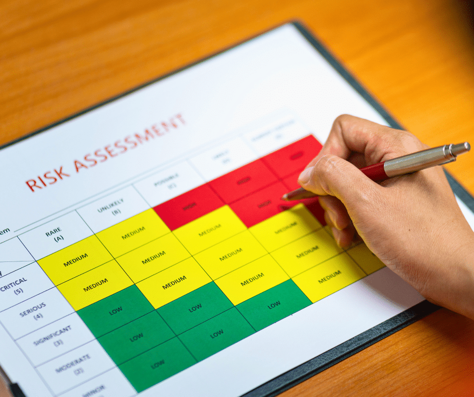 Artigo 2 Maio 2026 I - Vexillum Person marking a colored risk assessment chart labeled 'Risk Assessment' on a clipboard with a red pen. The chart uses red, yellow, and green blocks to categorize risk levels.