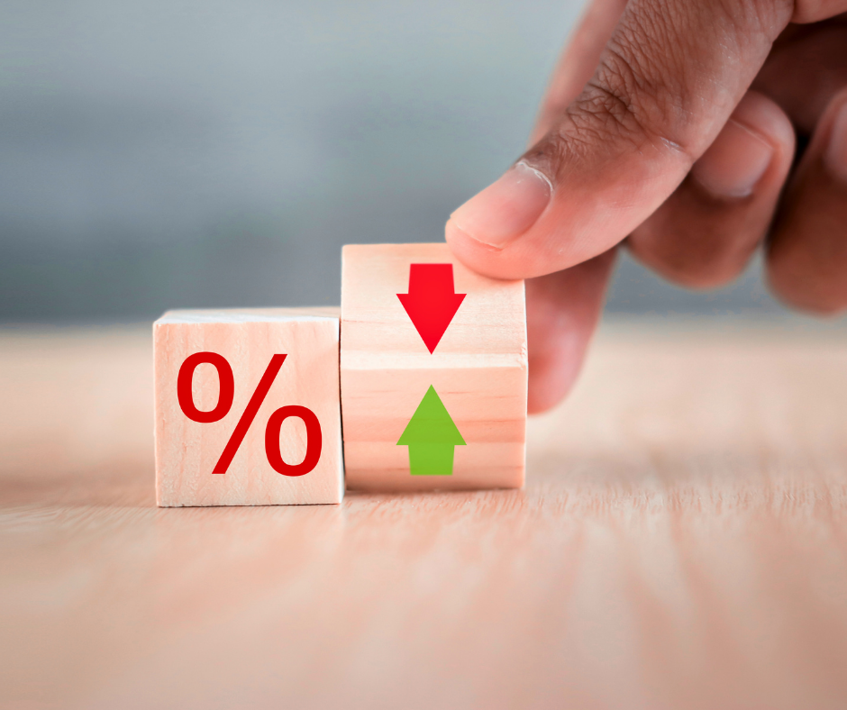 Artigo 4 Maio 2026 I - Vexillum Close-up of two wooden blocks: left block displays a red percent sign, right block shows a red downward arrow on top and a green upward arrow below, with a hand nudging the upper block, suggesting changing rates or prices.