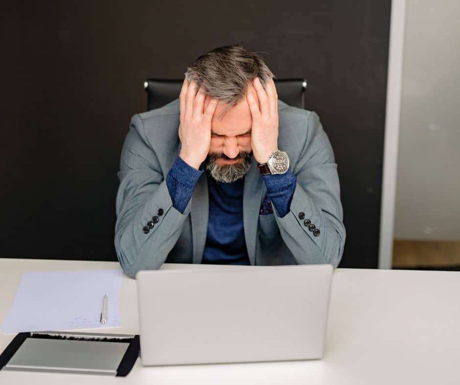 Frustrated businessman sits at a desk with a laptop, hands on his head in stress.